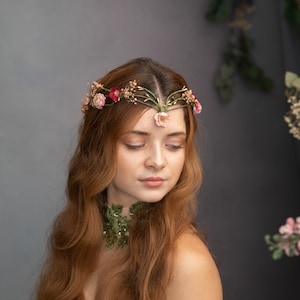 May include: A woman with long, wavy red hair wears a floral crown with pink and red flowers. She is wearing a green and white dress with a floral design. The background is a gray wall with greenery.