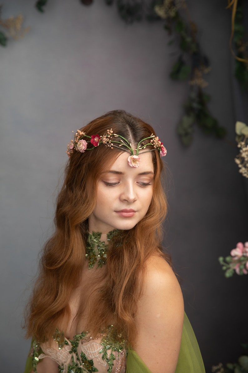 May include: A woman with long, wavy red hair wears a floral crown with pink and red flowers. She is wearing a green and white dress with a floral design. The background is a gray wall with greenery.