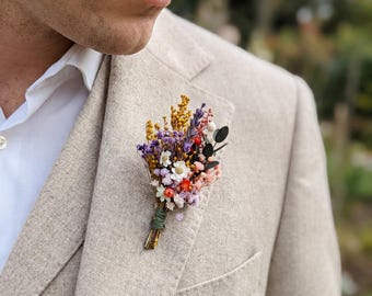 May include: A boutonniere of dried flowers in shades of yellow, purple, white, and pink, tied with green twine, pinned to a beige blazer. The blazer is worn over a white dress shirt. The image is a close-up.