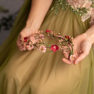 May include: A floral crown with pink and red flowers and green leaves. The crown is held in the hands of a person wearing a green dress.