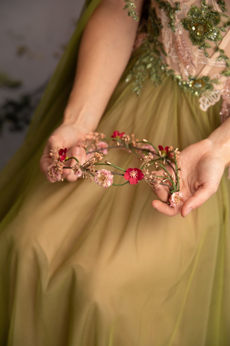 May include: A floral crown with pink and red flowers and green leaves. The crown is held in the hands of a person wearing a green dress.