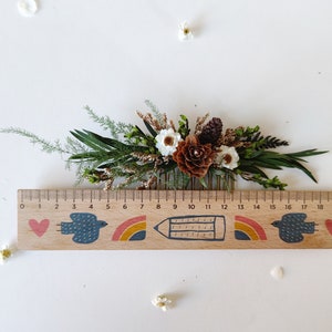 May include: A wooden comb with a floral arrangement of dried white flowers, brown pine cones, and green foliage. The comb is decorated with a painted design of a heart, a bird, a rainbow, a pencil, and another rainbow. The ruler below the comb is marked with numbers from 0 to 20.
