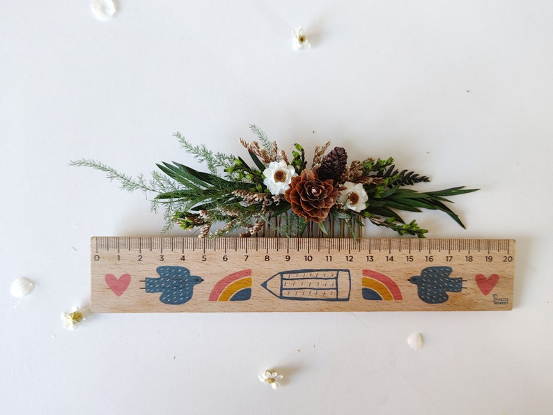 May include: A wooden comb with a floral arrangement of dried white flowers, brown pine cones, and green foliage. The comb is decorated with a painted design of a heart, a bird, a rainbow, a pencil, and another rainbow. The ruler below the comb is marked with numbers from 0 to 20.