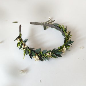 May include: A handmade floral crown with dark green leaves, tiny white flowers, and delicate green sprigs. The crown is arranged on a dark brown headband, creating a natural and elegant accessory. Ideal for weddings or special occasions.