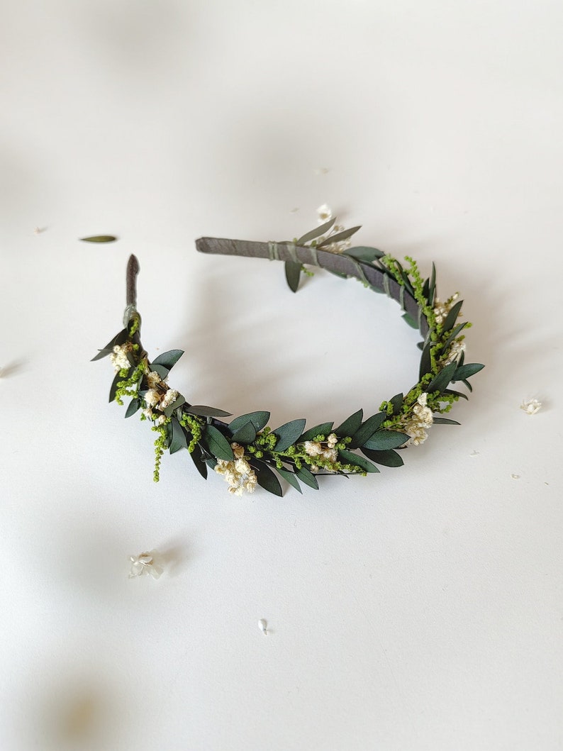 May include: A handmade floral crown with dark green leaves, tiny white flowers, and delicate green sprigs. The crown is arranged on a dark brown headband, creating a natural and elegant accessory. Ideal for weddings or special occasions.