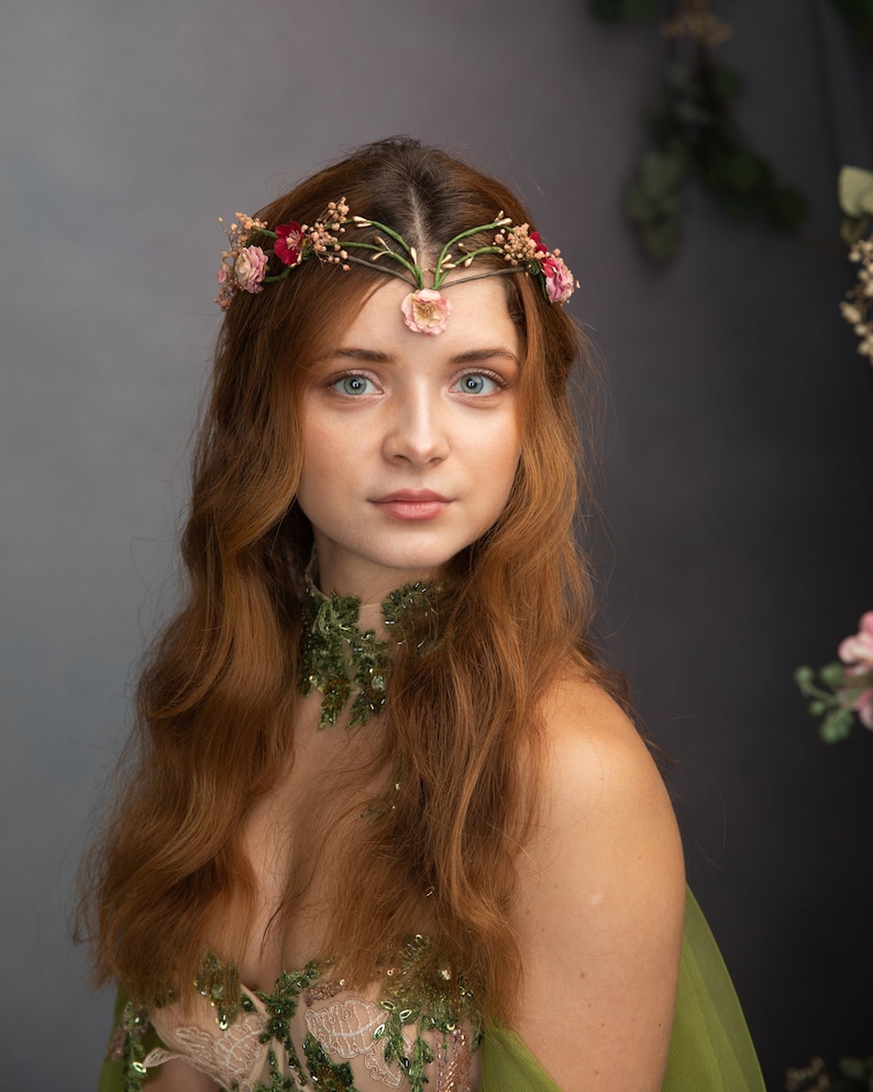 May include: A woman with long, red hair wearing a green and gold floral dress and a flower crown. She is looking directly at the camera.