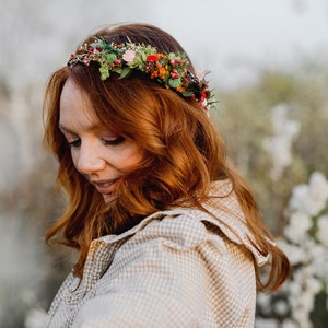 May include: A woman with long, red hair wears a floral crown with red, pink, and green flowers. She is wearing a light brown and white checkered shirt.