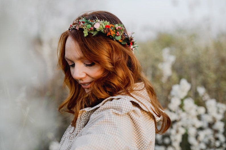 May include: A woman with long, red hair wears a floral crown with red, pink, and green flowers. She is wearing a light brown and white checkered shirt.