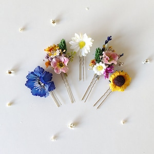 May include: Four floral hair pins with different flower arrangements. The pins feature a blue flower, a white daisy, a pink and purple flower arrangement, and a yellow sunflower.