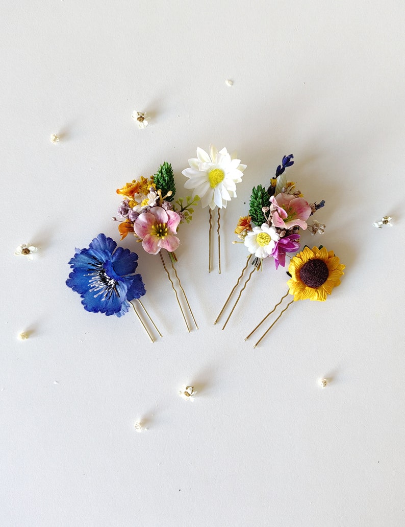 May include: Four floral hair pins with different flower arrangements. The pins feature a blue flower, a white daisy, a pink and purple flower arrangement, and a yellow sunflower.