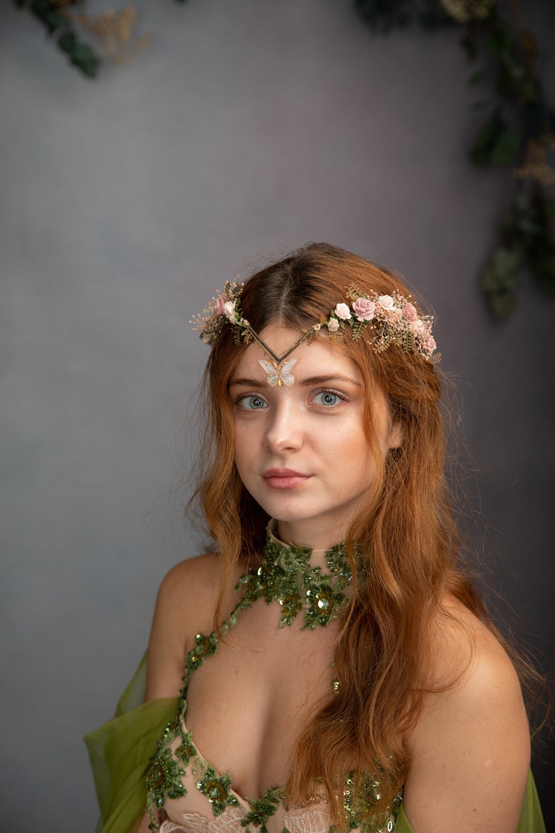 Peut inclure: Une femme aux longs cheveux roux portant une robe verte et beige avec une couronne de fleurs et une broche en forme de papillon. La robe a un col en sequins et une superposition transparente.