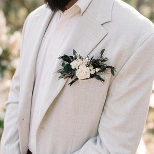 Puede incluir: Una blazer gris claro con una camisa blanca abotonada y un prendido floral. El prendido presenta rosas blancas, hojas verdes oscuras y peque&ntilde;as flores blancas. La blazer tiene un aspecto texturizado.