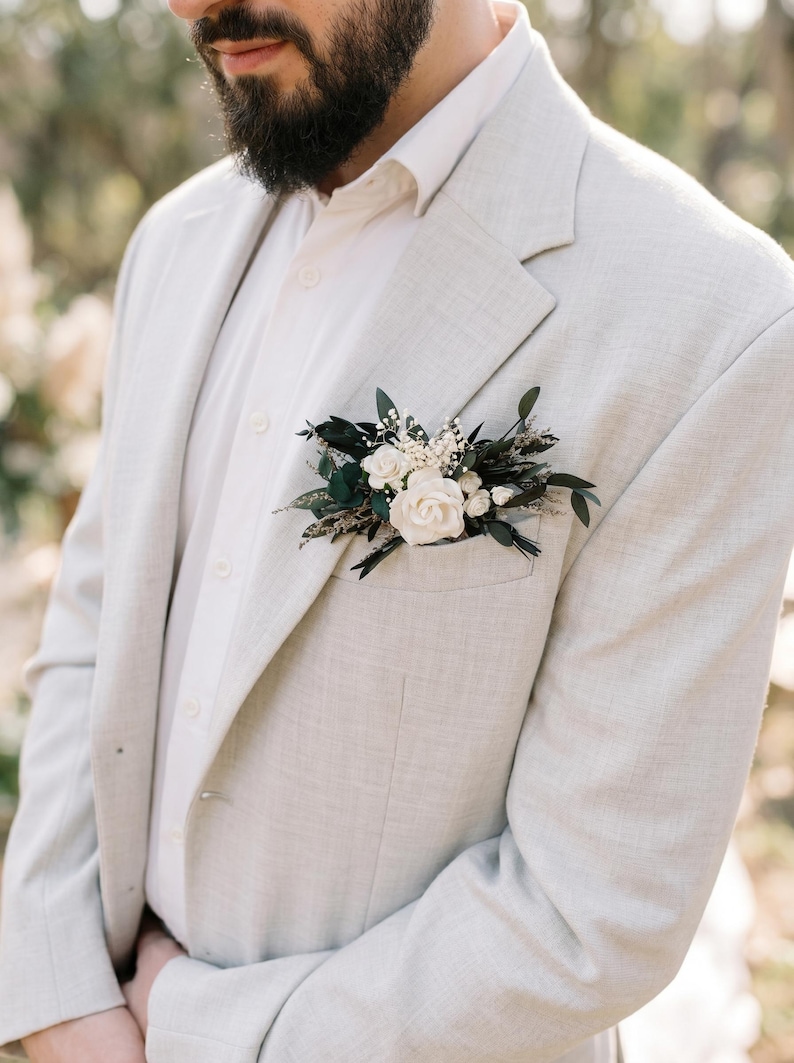 Puede incluir: Una blazer gris claro con una camisa blanca abotonada y un prendido floral. El prendido presenta rosas blancas, hojas verdes oscuras y peque&ntilde;as flores blancas. La blazer tiene un aspecto texturizado.