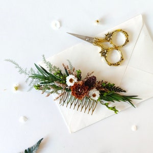 May include: A gold hair comb adorned with a floral arrangement of white and brown flowers, green leaves, and pine cones. The comb is on a white surface with a pair of gold scissors.