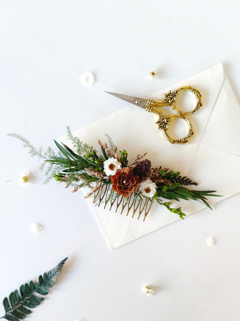 May include: A gold hair comb adorned with a floral arrangement of white and brown flowers, green leaves, and pine cones. The comb is on a white surface with a pair of gold scissors.