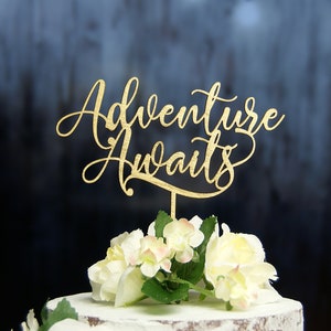 May include: Gold wooden cake topper with the text "Adventure Awaits" in a cursive font. The topper is placed on a white cake with white flowers.