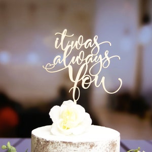 May include: A white cake with a wooden cake topper that says "it was always you". The cake topper is decorated with a small heart.