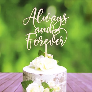 May include: A wooden cake topper with the words "Always and Forever" in cursive script with a heart symbol. The topper is on a white cake with white flowers.