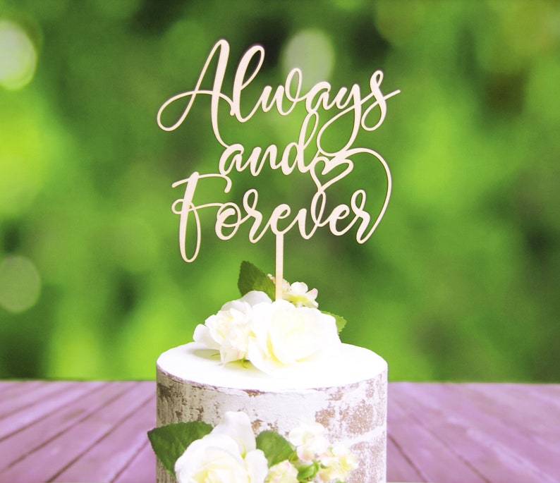 May include: A wooden cake topper with the words "Always and Forever" in cursive script with a heart symbol. The topper is on a white cake with white flowers.