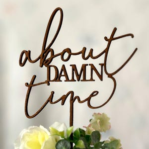 May include: Wooden cake topper with the text "about damn time" in a cursive font. The topper is brown and has a rustic look.