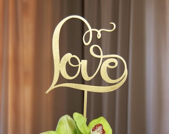 May include: A gold-colored cake topper in the shape of a heart with the word "love" in cursive script. The topper is placed above green orchids with red and white accents. The background is a blurred brown and beige.
