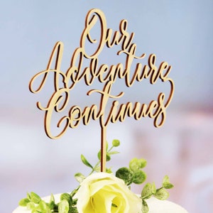 May include: Wooden cake topper with the text "Our Adventure Continues" in cursive script. The topper is on a white cake with green leaves and a white rose.