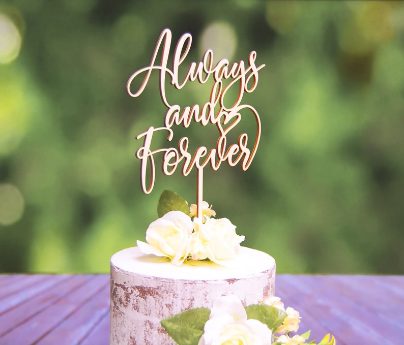 May include: Wooden cake topper with the words "Always and Forever" in cursive script with a heart symbol. The topper is on a white cake with white flowers.