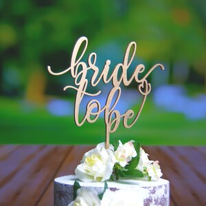 May include: A wooden cake topper with the words "bride to be" written in cursive script. The topper is placed on a white cake decorated with white flowers.