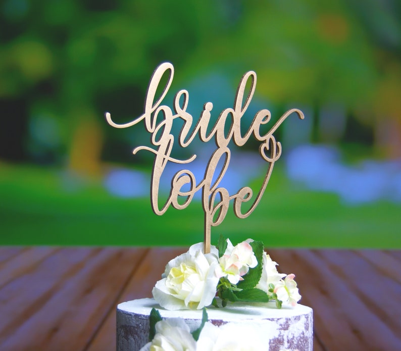 May include: A wooden cake topper with the words "bride to be" written in cursive script. The topper is placed on a white cake decorated with white flowers.