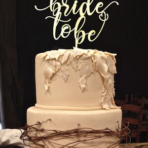 May include: A two-tiered wedding cake decorated with a brown vine and white leaves. The top tier has a gold "bride to be" cake topper.