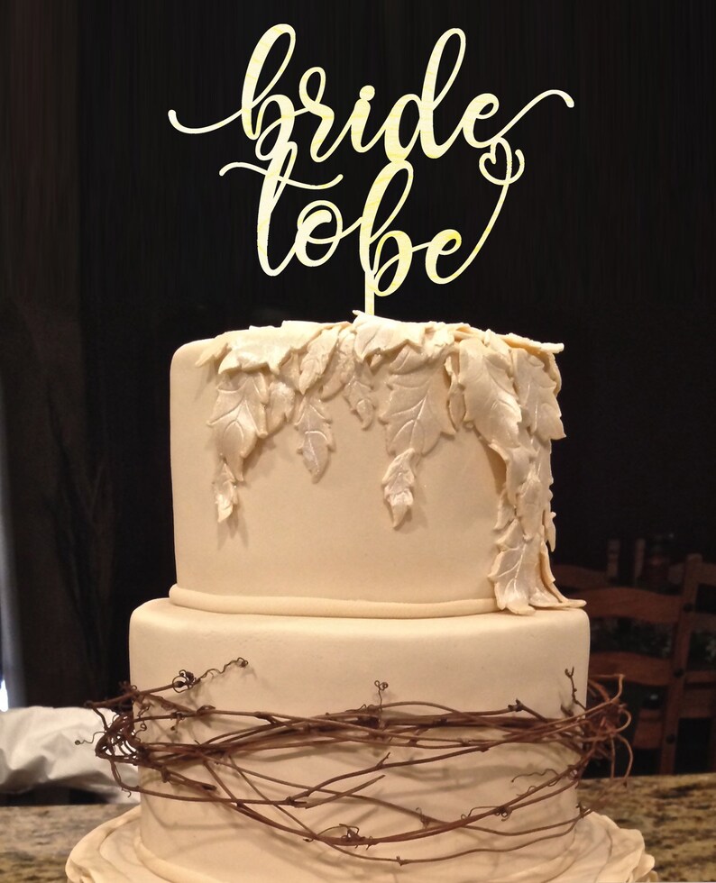 May include: A two-tiered wedding cake decorated with a brown vine and white leaves. The top tier has a gold "bride to be" cake topper.