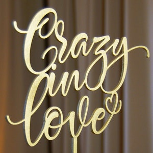 Pu&ograve; includere: Un cartello in legno dorato con le parole "Crazy in Love" scritte in corsivo. Il cartello ha una piccola forma di cuore alla fine della parola "Love".