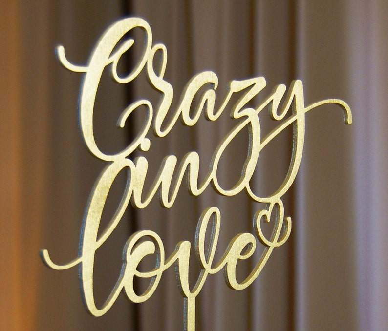 Pu&ograve; includere: Un cartello in legno dorato con le parole "Crazy in Love" scritte in corsivo. Il cartello ha una piccola forma di cuore alla fine della parola "Love".