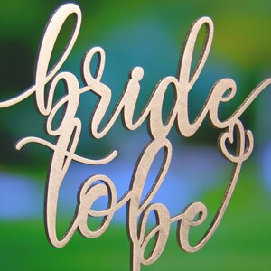 May include: Gold-colored wooden cake topper with the words "bride to be" in a cursive script.