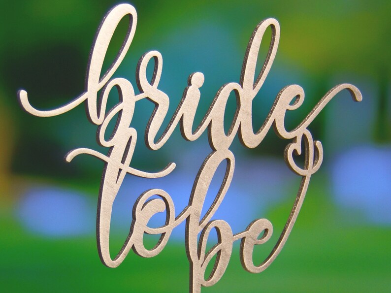 May include: Gold-colored wooden cake topper with the words "bride to be" in a cursive script.