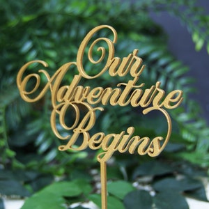 May include: Gold wooden sign with the words "Our Adventure Begins" in a cursive font.