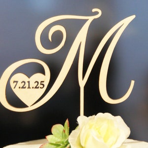 May include: Wooden cake topper with a cursive letter M and a heart with the date 7.21.25.