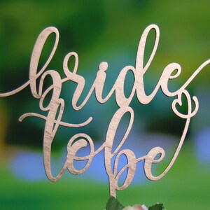 May include: A gold-colored wooden cake topper with the words "bride to be" written in a cursive font.