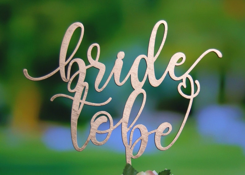 May include: A gold-colored wooden cake topper with the words "bride to be" written in a cursive font.
