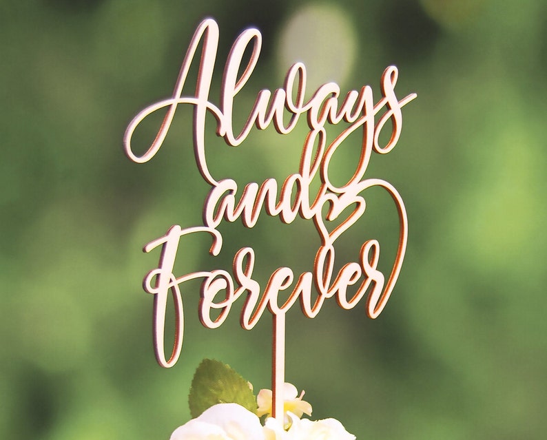 May include: Wooden cake topper with the words "Always and Forever" in a cursive font. The topper is a light brown color and has a heart shape in the word "and".