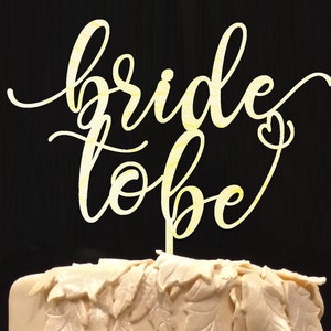 May include: A gold-colored wooden cake topper with the words "bride to be" written in a cursive font.