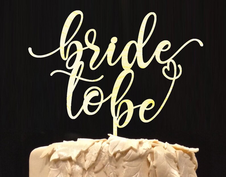 May include: A gold-colored wooden cake topper with the words "bride to be" written in a cursive font.