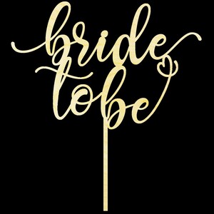 May include: A wooden cake topper with the words "bride to be" written in a cursive font. The topper is a light brown color and has a small heart at the end of the "be".