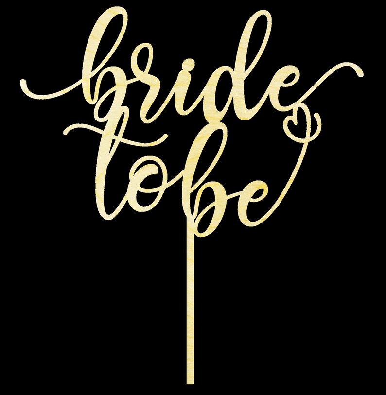 May include: A wooden cake topper with the words "bride to be" written in a cursive font. The topper is a light brown color and has a small heart at the end of the "be".