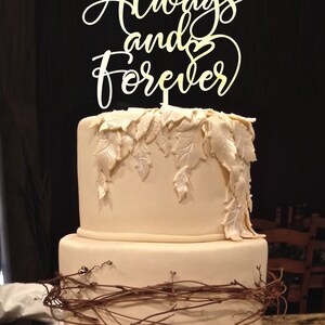 May include: A two-tiered wedding cake decorated with brown vines and white leaves. The cake topper is a gold sign that reads "Always and Forever" with a heart.