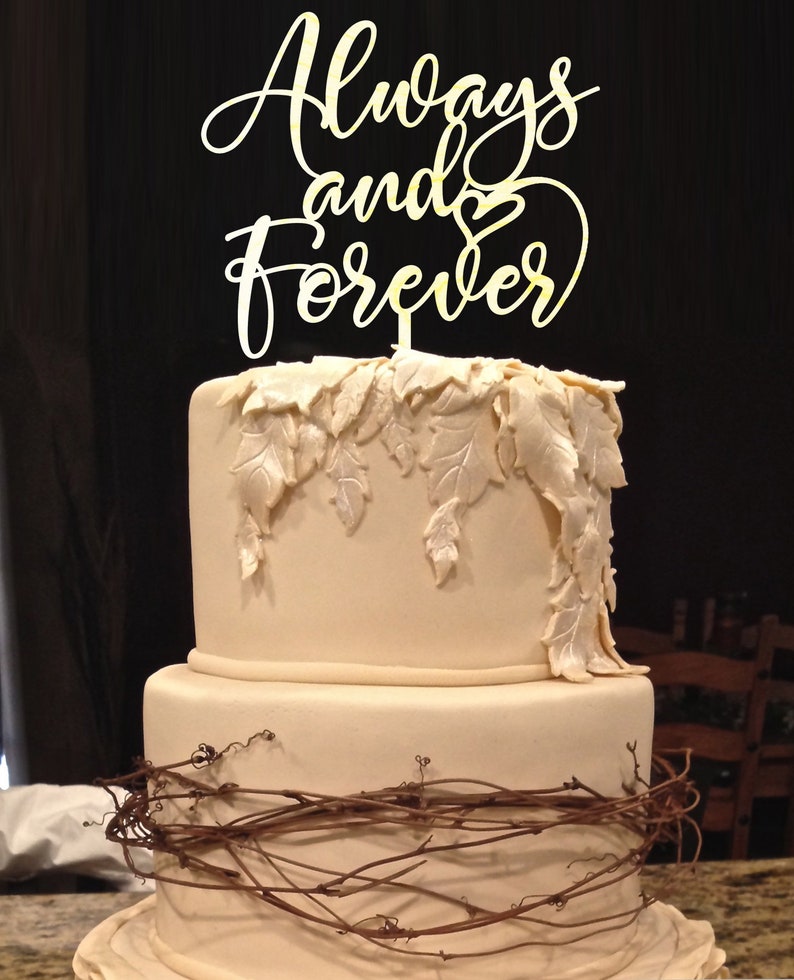 May include: A two-tiered wedding cake decorated with brown vines and white leaves. The cake topper is a gold sign that reads "Always and Forever" with a heart.