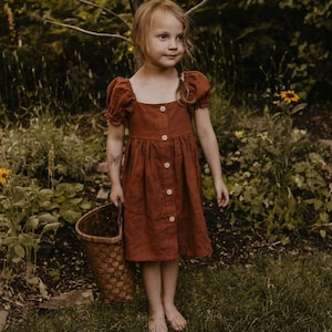 Puede incluir: Una niña pequeña con un vestido de lino color óxido con mangas abullonadas y cierre de botones. Lleva una cesta tejida y está de pie en un jardín.