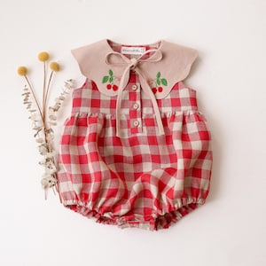 May include: A red and beige plaid baby romper with a beige collar. The collar is adorned with embroidered cherries and a bow. The romper has three buttons down the front and elastic around the legs.