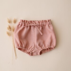 May include: Dusty pink bloomers featuring an elasticated waistband with a decorative drawstring tie and elasticated leg openings. The garment has a textured fabric appearance and is displayed alongside a small cluster of dried grass stems.