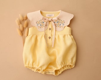 May include: A pale yellow romper featuring a cream-colored scalloped collar with delicate floral embroidery. The garment includes a front button placket with two visible buttons, a tie closure at the neckline, and a label reading DANNE & LOU.
